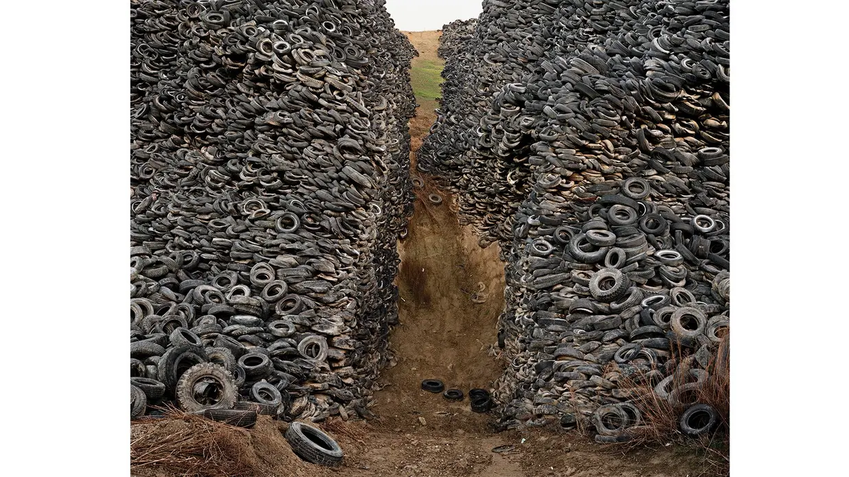 عکس تودهٔ عظیمی از لاستیک‌های دور ریخته که به وسیلهٔ مسیر خاکی در وسط تقسیم شده و تپهٔ سبزی در پس‌زمینه به‌دست می‌آید