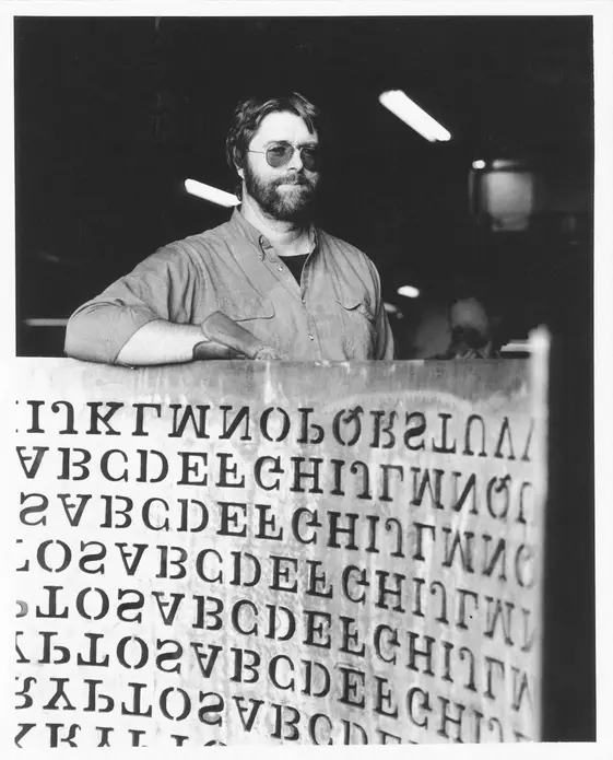 مجسمه‌ساز، جیم سنبورن، کنار مجسمهٔ مسی «کریپتوس» خود که نوشته‌هایی از حروف روی آن حک شده است.