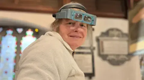 زنی با موهای خاکستری که دستگاه بزرگنمایی بر سر دارد، نیمهلبخند به دوربین میدهد. او یک پوشاک گرم و پشمی به تن دارد و میتوانید شیشههای رنگی و تابلوهای کلیسا را در پسزمینه مشاهده کنید.