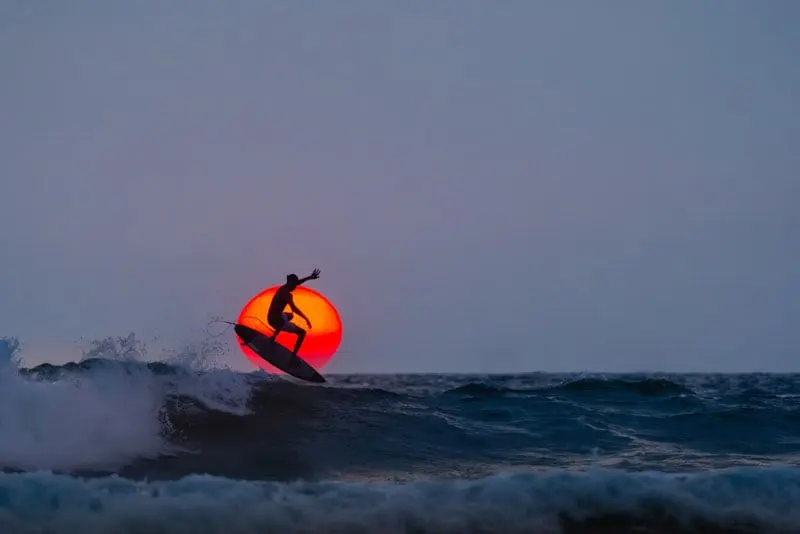 سورفرِی بر روی موجی در غروب سوار می‌شود؛ به‌صورت سیلوئهٔ تاریک در برابر خورشید نارنجی که پایین افق قرار دارد، با امواج تیره و آسمانی صاف در پس‌زمینه.