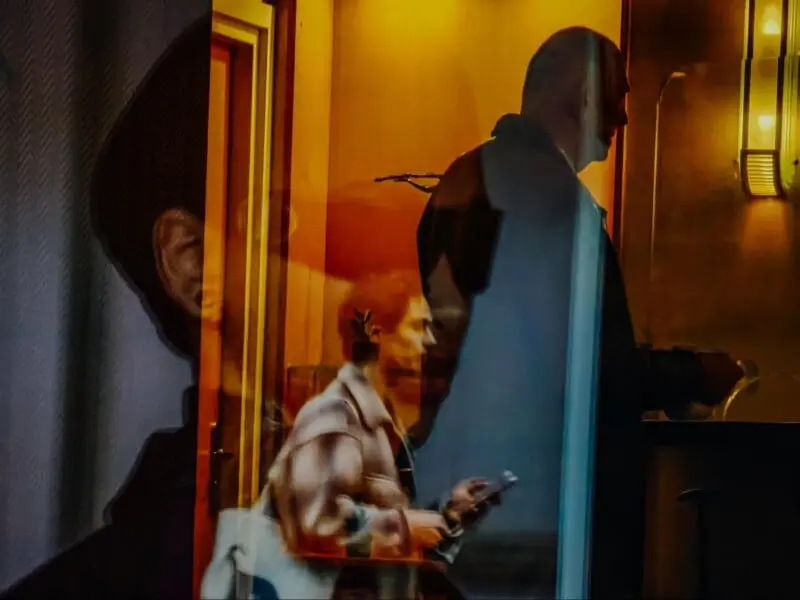 عکسی لایهدار و انتزاعی که سایهها و بازتاب افراد را در نور گرم داخلی نشان میدهد؛ یک نفر در حال راه رفتن با تلفن، دیگری ایستاده در پیشخوان، هر دو بهنحوی همپوشانی دارند.